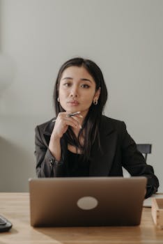 Asian woman in business attire working on a laptop at home, focusing on an online task.