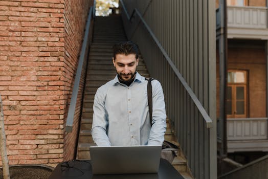 Businessman working remotely on a laptop outdoors by brick wall and stairs.